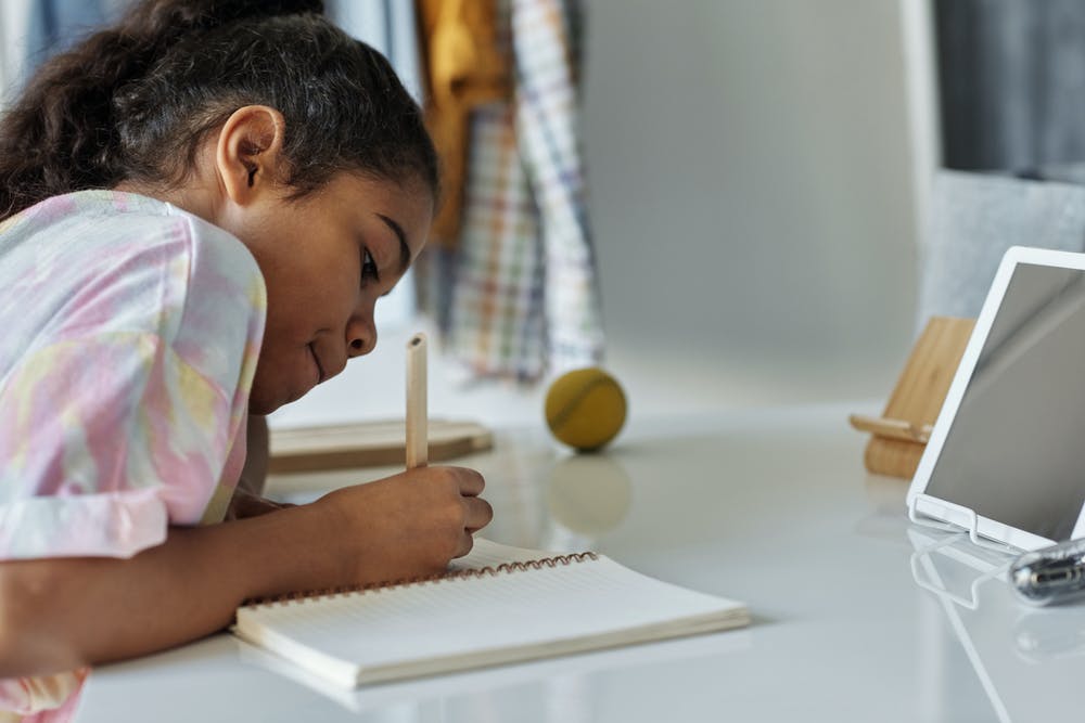 A young woman wearing tie-dye leans over to write dutifully in her notebook, as if recording something she saw or heard on the screen in front of her