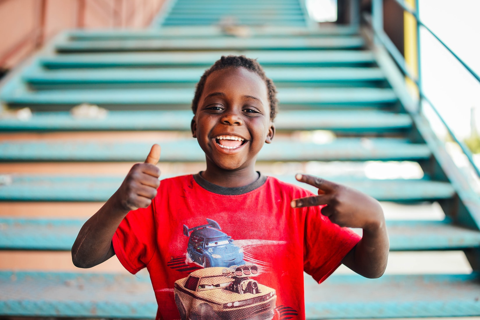 A young boy, wearing a red Cars T shirt and a big smile, stands in front of a turquoise outdoor staircase. He gives a thumbs up with one hand and a peace sign with the other