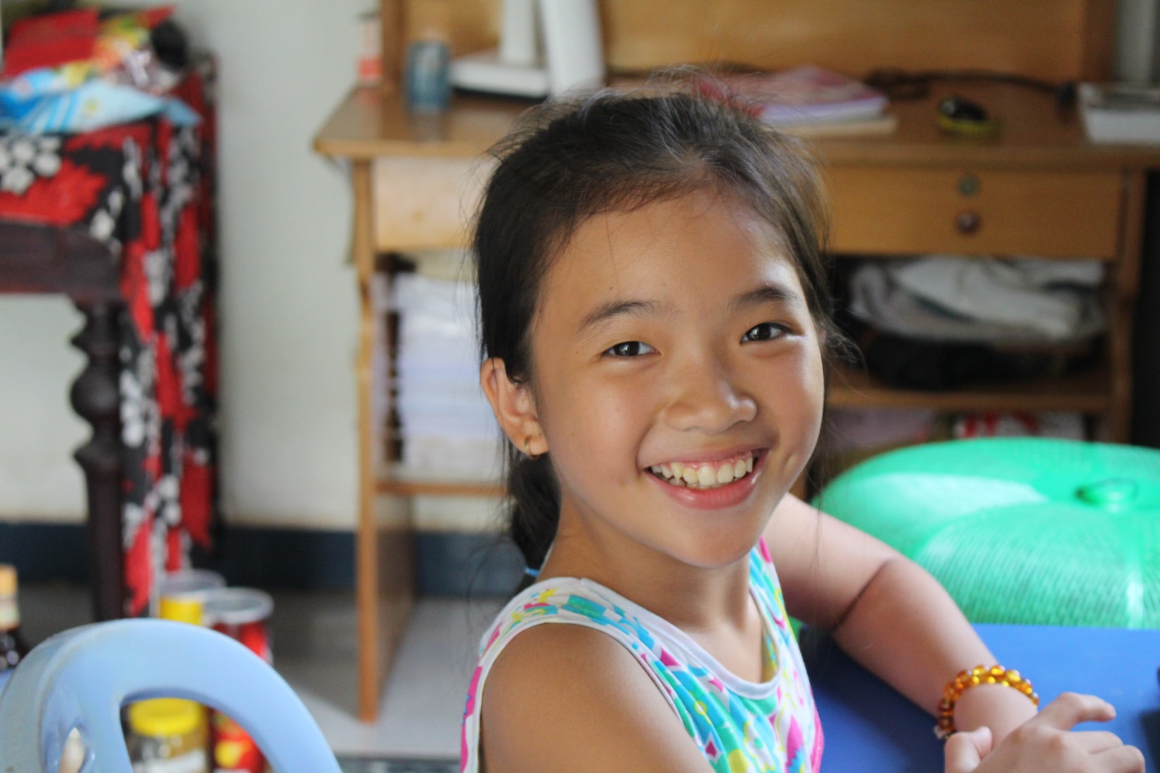 A young girl smiles toward the camera - she is in a multicolored tank top and seated comfortably in a common area